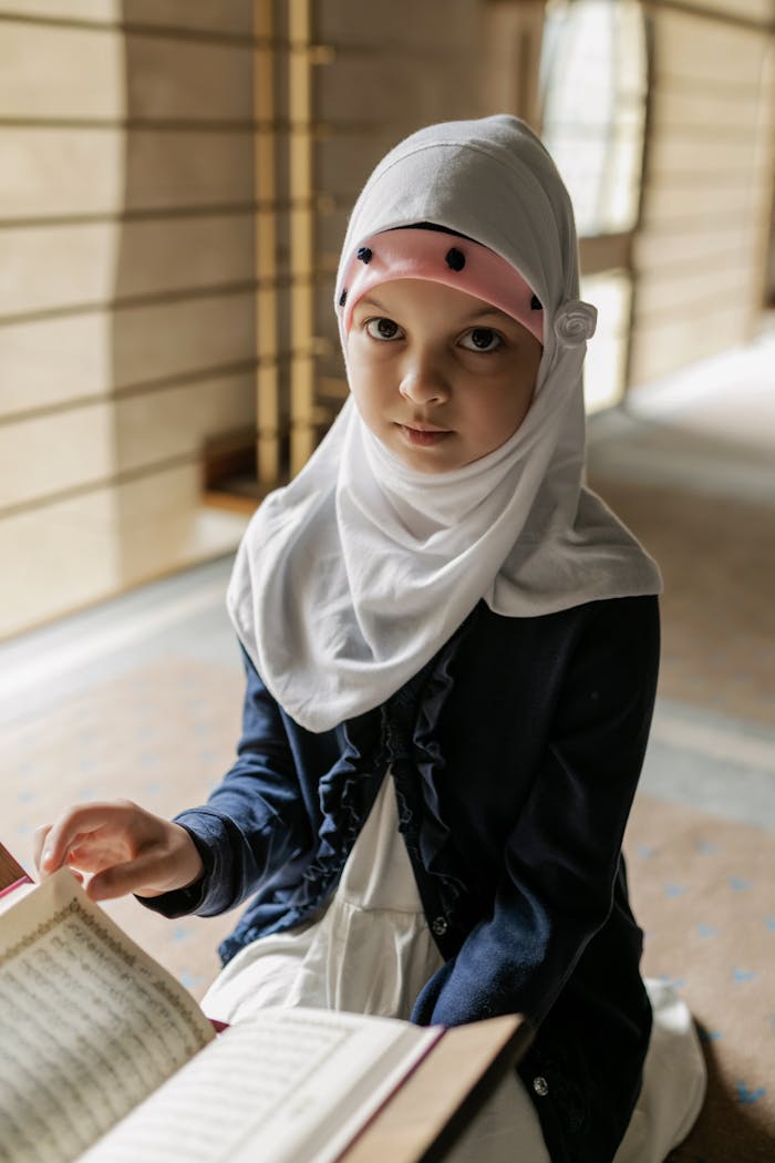 services-03 Portrait of a young girl in a hijab reading the Quran indoors. The scene captures a peaceful, cultural moment.